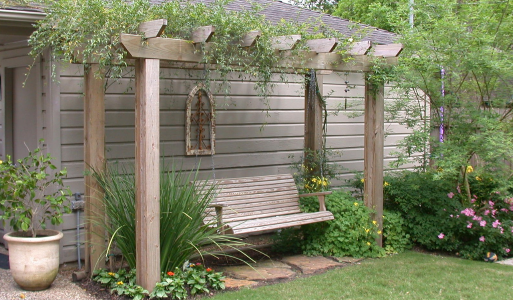 Wood pergola with climbing greenery and a hanging bench surrounded by landscaped garden beds and lawn.