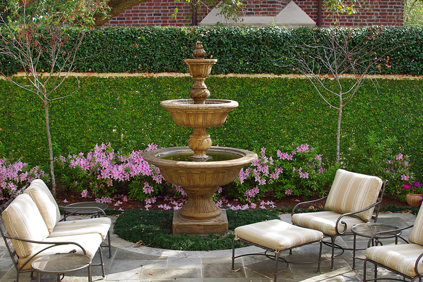 Three tiered fountain surrounded by pink flowers and iron outdoor seating.